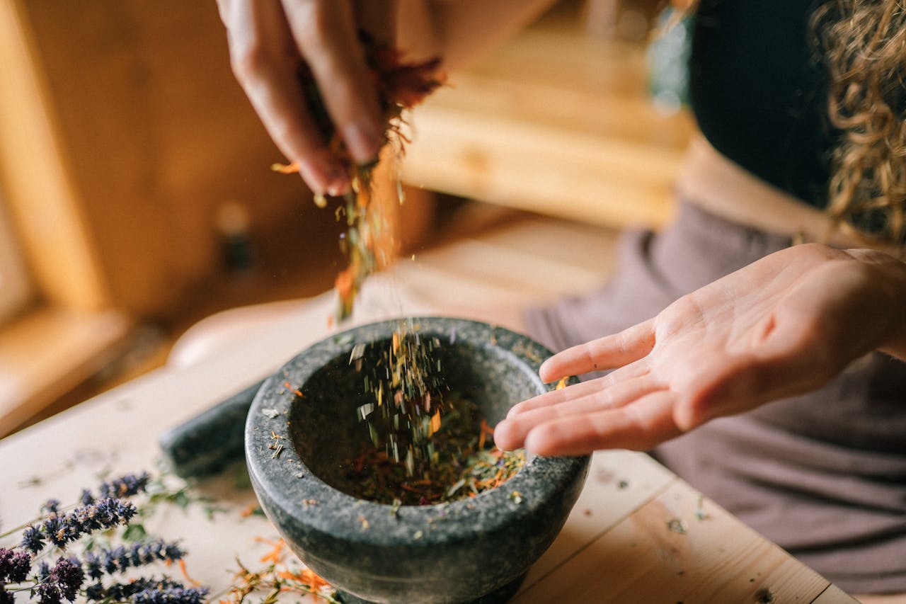 A person preparing dried herbs using a mortar and pestle on a wooden table, seen from above.